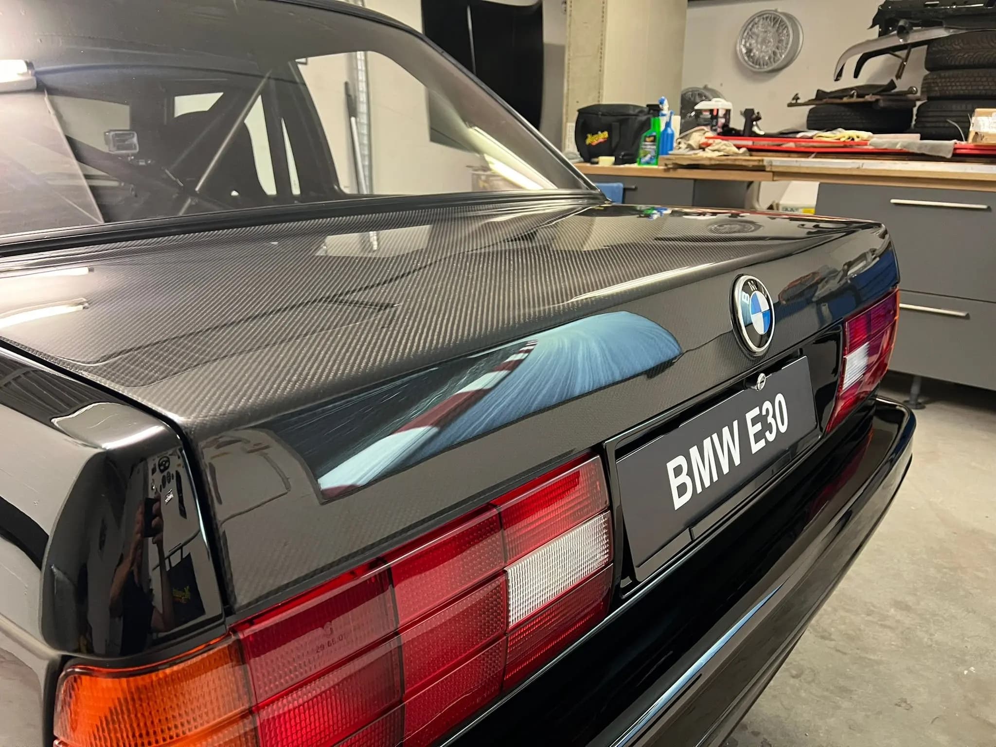 Eingebauter Sichtcarbon-Kofferraumdeckel am BMW E30 in der Werkstatt, Heckansicht mit Rückleuchten und BMW-Emblem.
