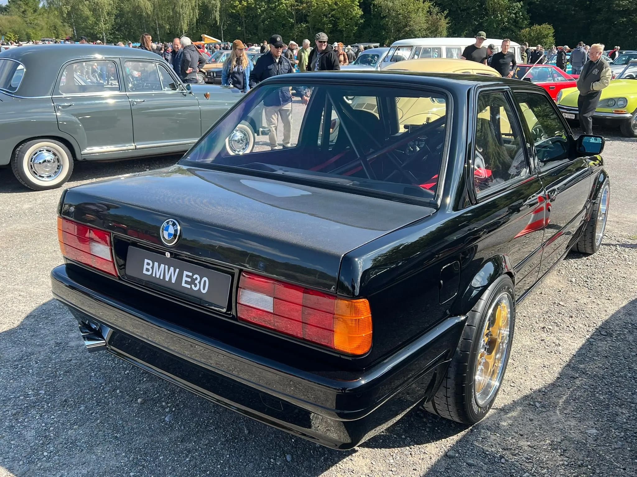 Schwarzer BMW E30 von hinten schräg auf einem Treffen, mit Carbon-Kofferraumdeckel und breiten Rädern.