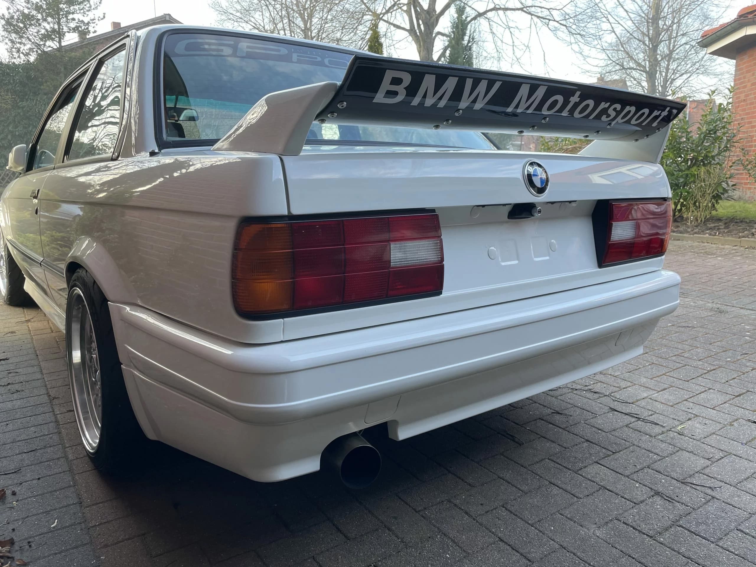 Heckansicht schräg: weißer BMW E30 mit großem BMW Motorsport Heckflügel.