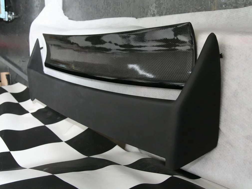BMW E30 M3 Heckspoiler-Set schräg aufgestellt: matte Basis mit Endkappen und glänzendem Sichtcarbon-Einsatz dahinter.