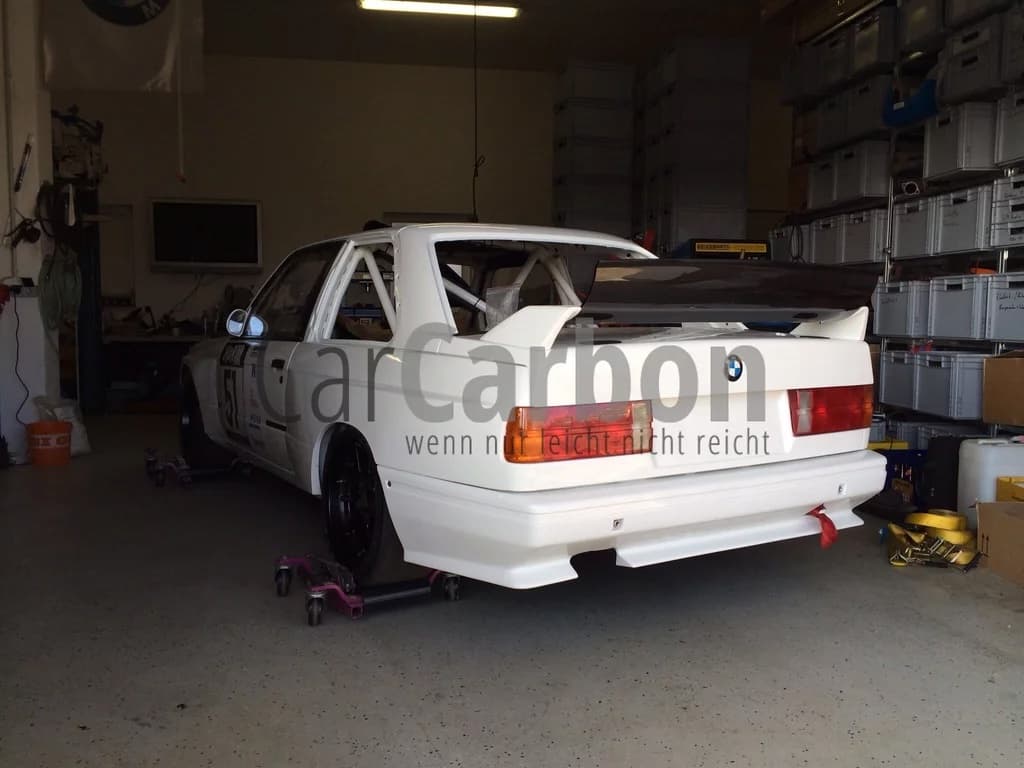 BMW E30 M3 von hinten in der Werkstatt, montierte weiße Heckschürze mit Carbonleiste und Abschleppgurt.